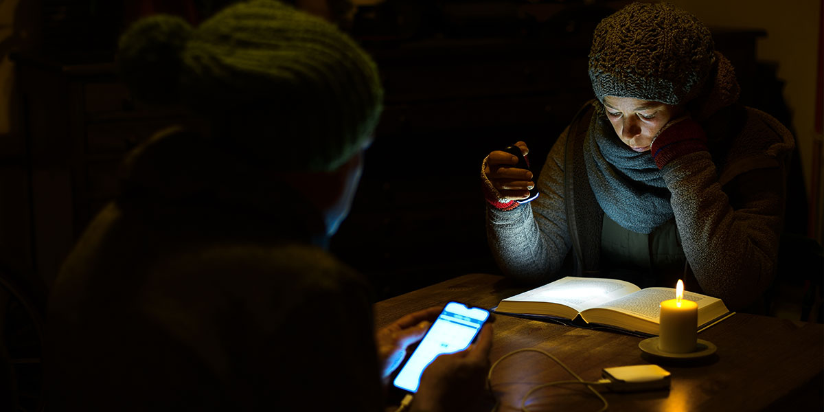 Woman reading a book by torchlight and woman using a phone connected to a powerbank in a dark room